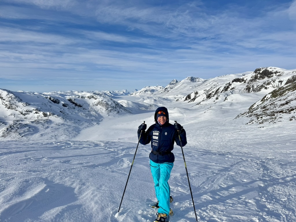 Blide Kristine går på truger på det snøkledde fjell.
Hun har på seg en mørkeblå jakke med hette, oransje solbriller og lyseblå vinterbukse.
