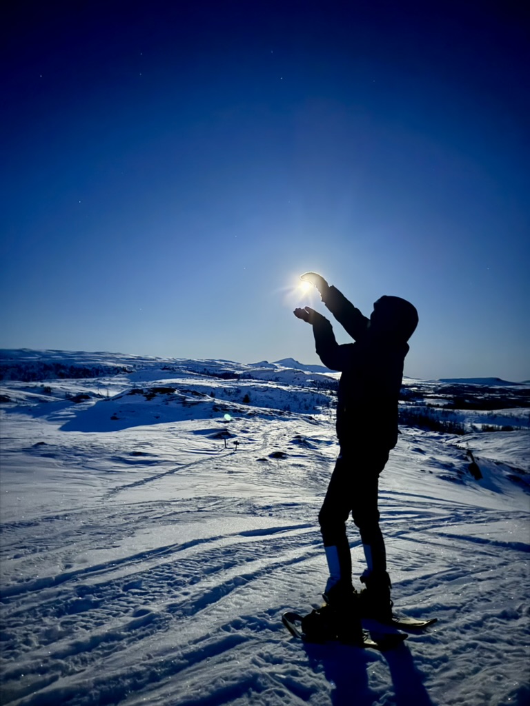 man ser kontorene av Kristine med en sterk fjellsol i bakgrunnen. Hun har på seg truger i et snøkledd fjellandskap. Hun holder hendene rundt solen, så det ser ut som hun har fanget den.