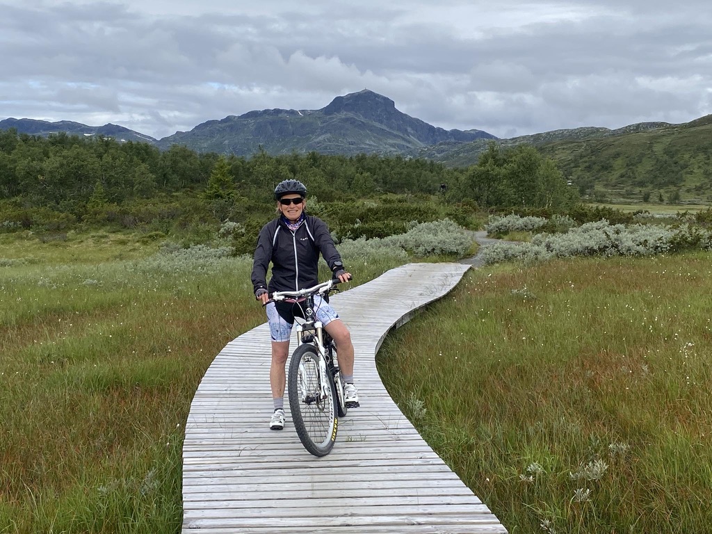 Kristine som sykler på en trebru over en myr. Bak henne er en sti som fører inn i fjellet. Bak henne er det skog og lengst bak i horisonten er fjellet.