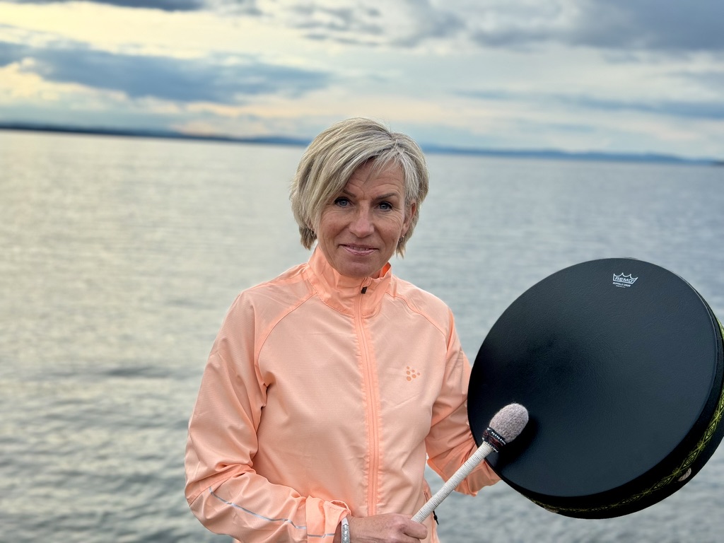 Kristine med sjamantromme. Hun er kledd i pastelloransje jakke og i bakgrunnen er havet, med overskyet himmel.
