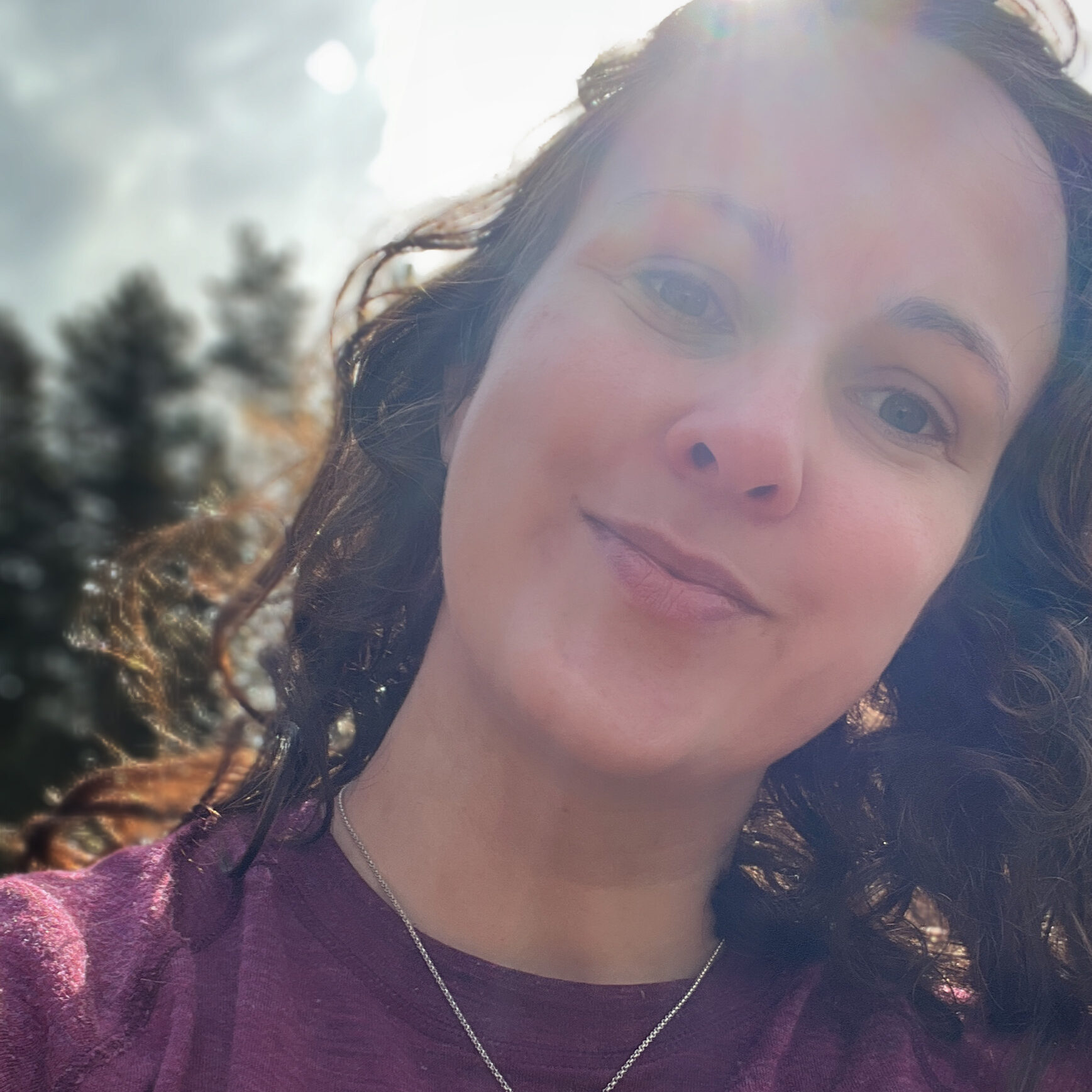 Selfie av Siv i lilla ulltrøye med solstråler og skogen i bakgrunn.