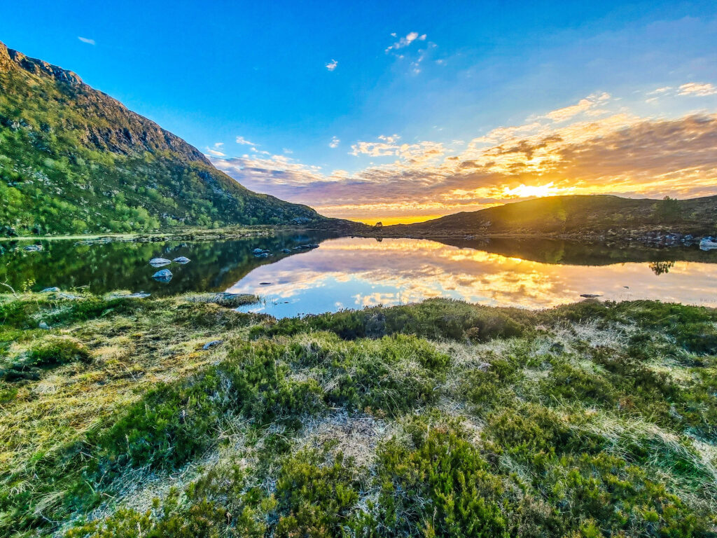 Solnedgangen reflekteres i vannet ved Stordalsvatnet, omgitt av grønn natur i et fjellandskap.