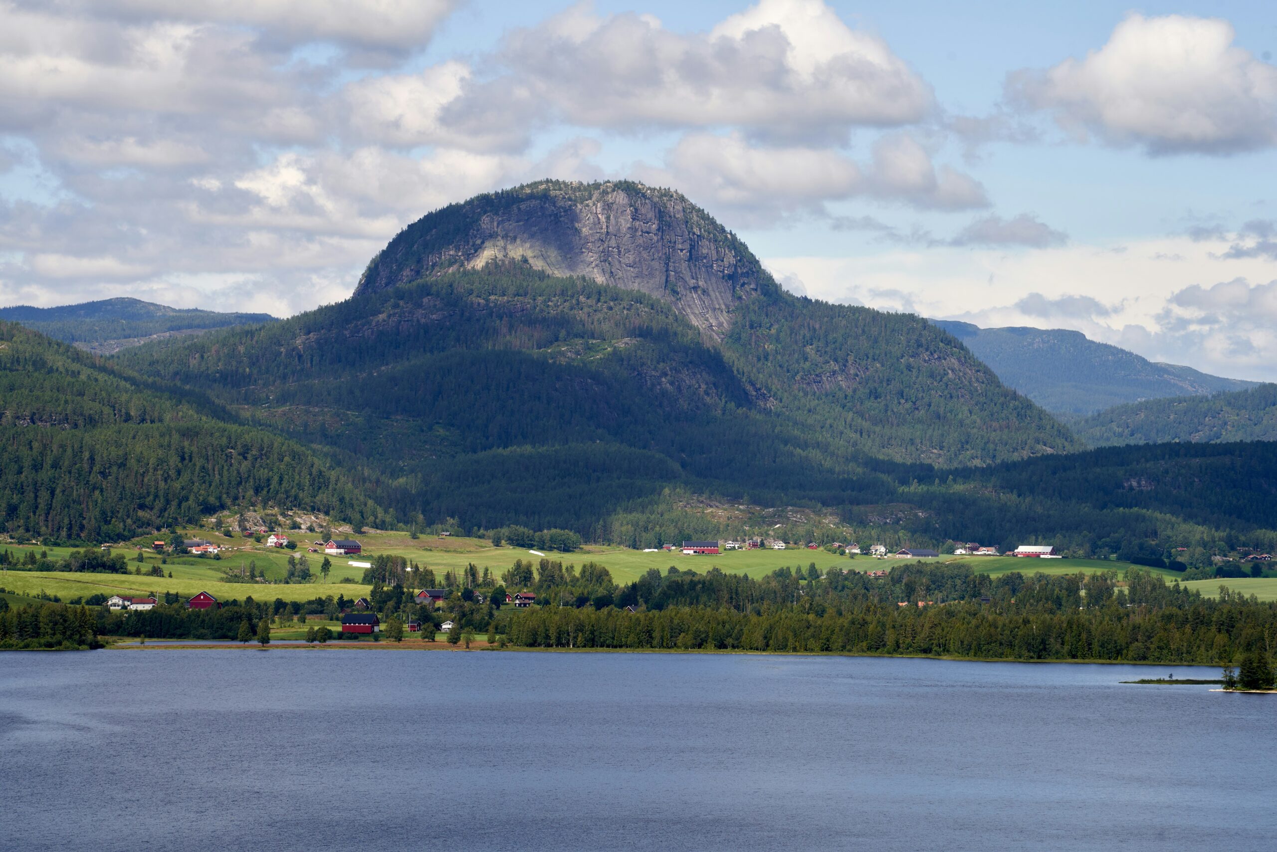 Andersnatten i Sigdal kommune. Omgitt av skog. Blått, stille vann i forgrunnen under en blå lett overskyet himmel.