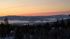 Solnedgang over snøkledd dal, dekt med tåke og skog.