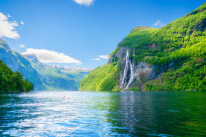 De syv søstre fossene i Geirangerfjorden. Over speilblankt mann, majestetiske grønnkledde fjell under en blå himmel.