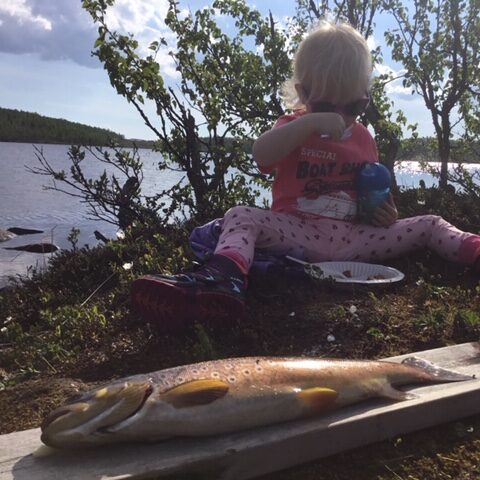 Lite barn spiser mat ute i det fri. Foran ligger en nyfisket fisk på ei fjøl.
