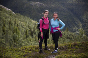 To sportykledde, smilende turdamer i et grønt og skogkledd landskap.