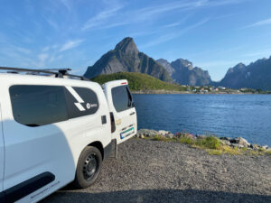 Selvbygd campingbil i Lofoten med Reine i bakgrunn, under en blå himmel.