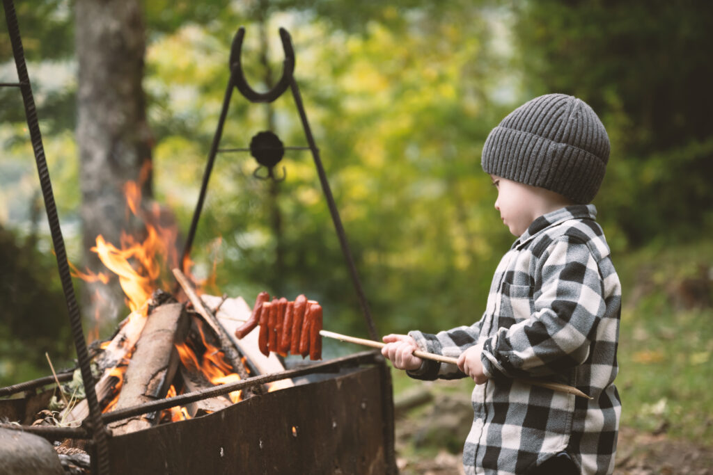 Lite barn i rutete skjorte og grå lye, griller pølse over et leirbål.