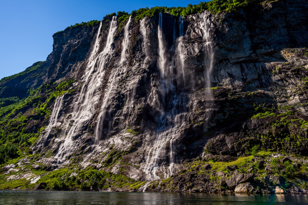 De syv søstre fossefallene i Geirangerfjorden i solskinn under en blå himmel, over stille vann. 