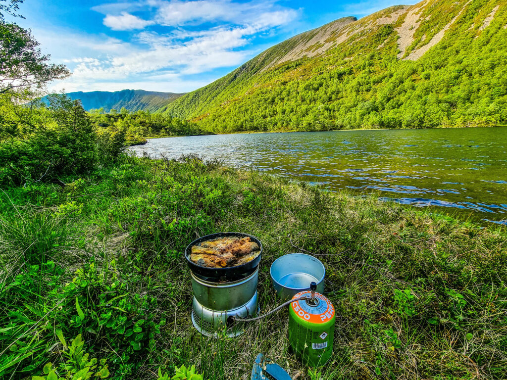 Matlaging i stormkjøkken. Deilig mat putrer i en svart panne over et stormkjøkken mellom lyng og gress. Gass er koblet til og en tom gryte/skål står ved sien av. I bakgrunnen renner en elv mellom grønne åser under en blå, lett overskyet himmel.