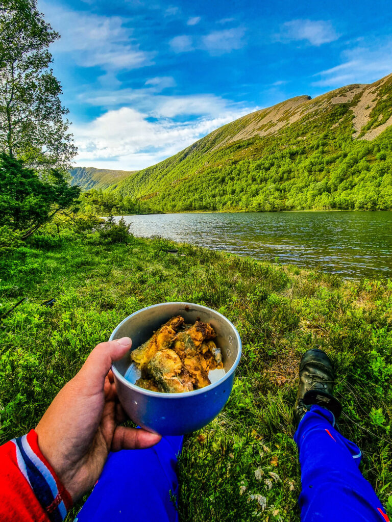 FjellTider Magnar holder nylaget middag i hånda, med grønn og frodige åser og rennende elv i bakgrunn.