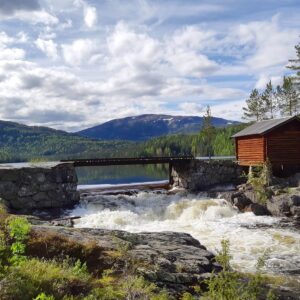 Naturlandskap ved buvasskoia. En enkel trebru over foss. Omgitt av grønn natur og rød koie på høyre side av brua. Blå skogkledde åser i bakgrunn. Under blå, overskyet himmel.
