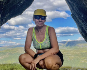 Marianne iført gul caps og turtøy for sommervær, sitter på huk mellom to store steiner. Med blå himmel, lett overskyet og vakkert, grønt fjellandskap i bakgrunn.