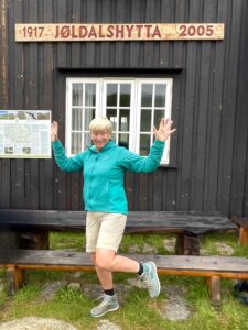 FjellTider Kari poserer foran tyinstølen. Veggen bak er brun med skiltet "tyinstålen" på et treskilt med rødmalte bokstaver.