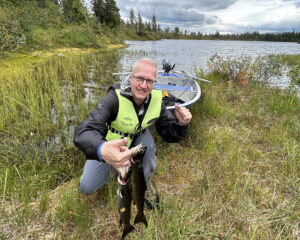 FjellTider Tomas triumferer med fiskefangsten. Foran båten ved et lite skogsvann.