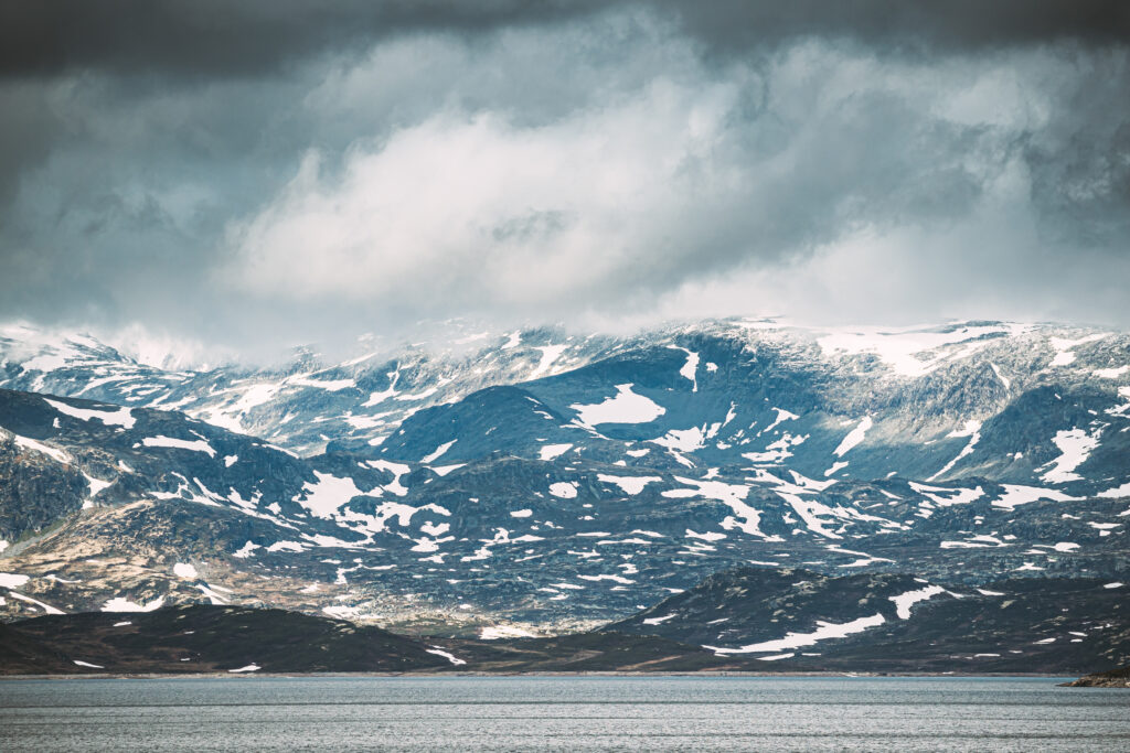 Tyin i Jotunheimen. Vann omgitt av høye fjell. Overskyet himmel og lett sollys.